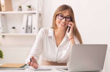 Ofisteki neşeli kız cep telefonuyla konuşuyor. Çalışma masasında oturuyor.