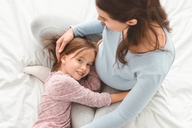 Little girl hugging pregnant moms belly while lying on bed clipart