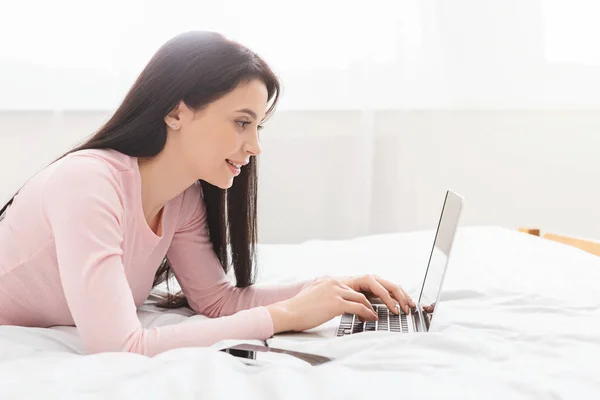 Millennial estudiante chica escribiendo en el ordenador portátil, acostado en la cama —  Fotos de Stock