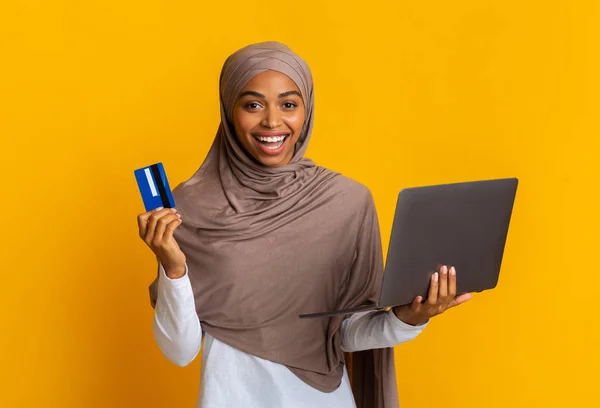 Радісна чорношкіра мусульманка в хіджабі тримає ноутбук і кредитну картку — стокове фото
