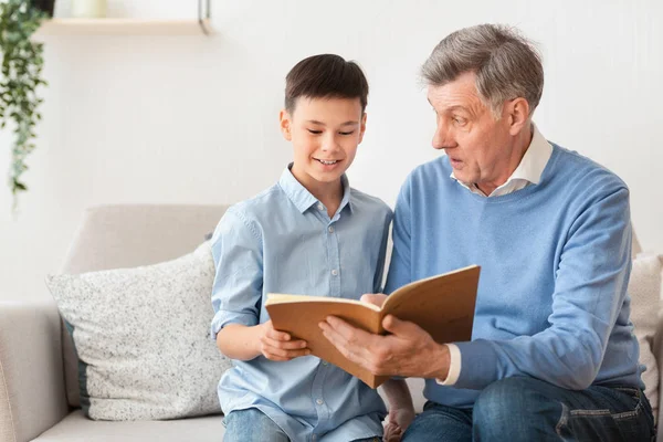 Дідусь і онук читають книгу сидячи на дивані вдома — стокове фото