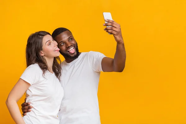 Радостная многорасовая пара делает селфи на смартфоне, гримасит и показывает языки — стоковое фото