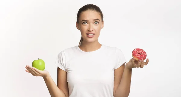 AppleとDoughnutの間で白い背景を選ぶ混乱した女の子 — ストック写真