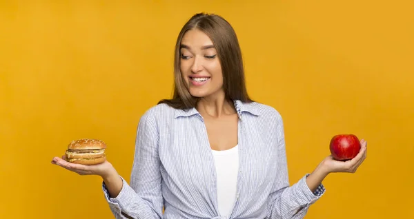 Girl Choosing Burger Instead Of Apple Standing On Yellow Background Stock Image