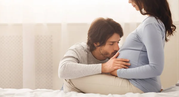 Orgulloso marido besando a sus esposas embarazadas vientre en la cama en casa —  Fotos de Stock