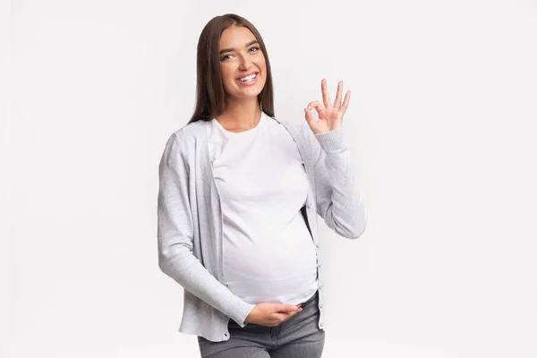 Feliz mulher grávida Gesturing Ok em pé sobre fundo branco — Fotografia de Stock