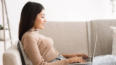 Genç bir kadın dizüstü bilgisayarda internette sörf yapıyor, evdeki kanepede dinleniyor.