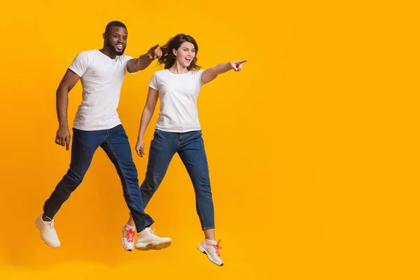 Saltando casal interracial apontando para espaço de cópia sobre fundo amarelo — Fotografia de Stock