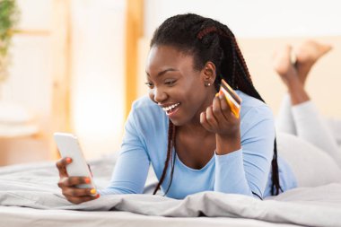 Akıllı telefon ve kredi kartı kullanan neşeli bayan yatakta uzanıyor.