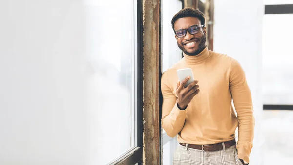 Χαμογελώντας μαύρο επιχειρηματία στέκεται κοντά στο παράθυρο με κινητό — Φωτογραφία Αρχείου