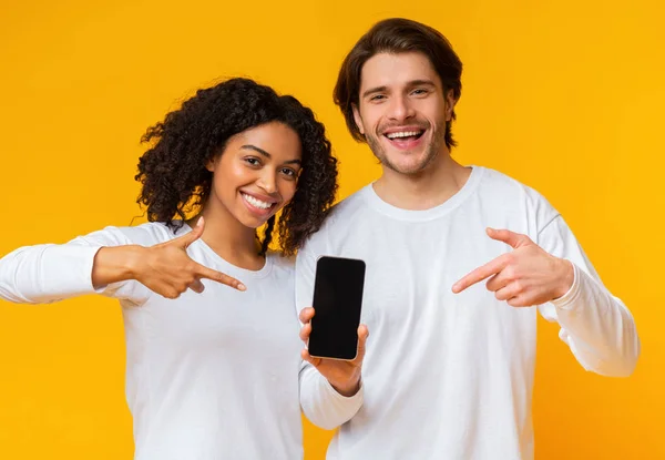 Pasangan antar ras ceria yang menunjuk pada smartphone dengan layar hitam untuk mockup — Stok Foto