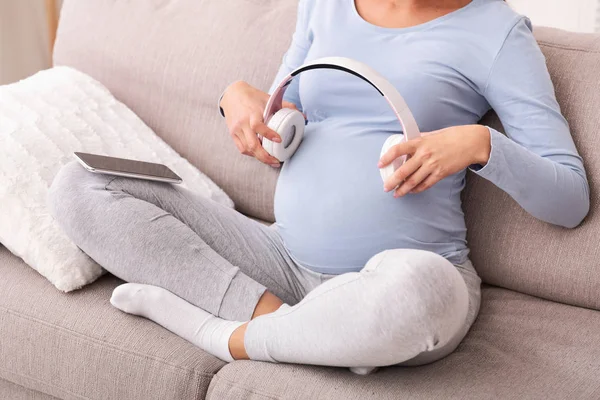 Mulher grávida irreconhecível segurando fones de ouvido perto da barriga sentado no sofá — Fotografia de Stock