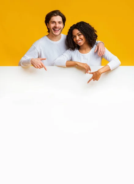 Joyful interracial couple leaning and pointing at white advertisement board — Stock Photo, Image