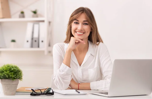 Счастливая деловая женщина сидит за ноутбуком улыбаясь в современном офисе — стоковое фото