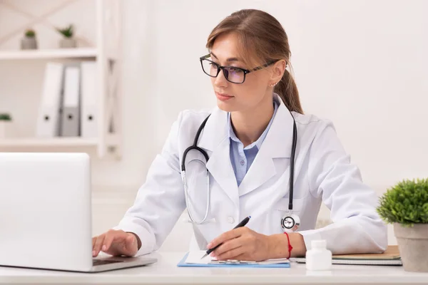 Doctora mujer trabajando en computadora portátil tomando notas sentada en la oficina —  Fotos de Stock