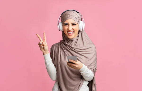 Gadis muslim yang ceria dengan headphone dan jilbab mendengarkan musik di smartphone — Stok Foto