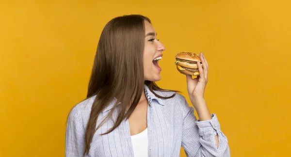 Ragazza felice mangiare hamburger malsano in piedi su sfondo giallo, Panorama — Foto Stock