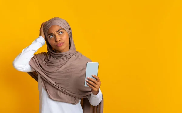 Pensativo afro musulmana chica sosteniendo teléfono inteligente y tocando su cabeza —  Fotos de Stock