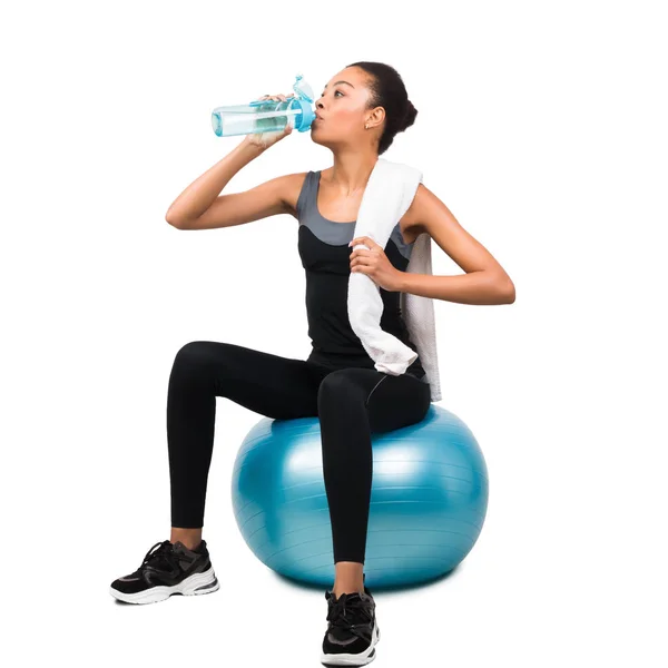 Afrikanische Amerikanerin trinkt Wasser sitzend auf Fitball, weißer Hintergrund — Stockfoto