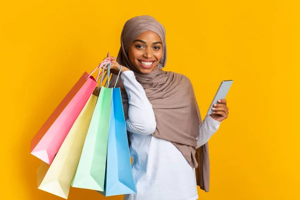 Akıllı telefon ve parlak alışveriş torbalarıyla poz veren neşeli Afro Müslüman kız. — Stok fotoğraf