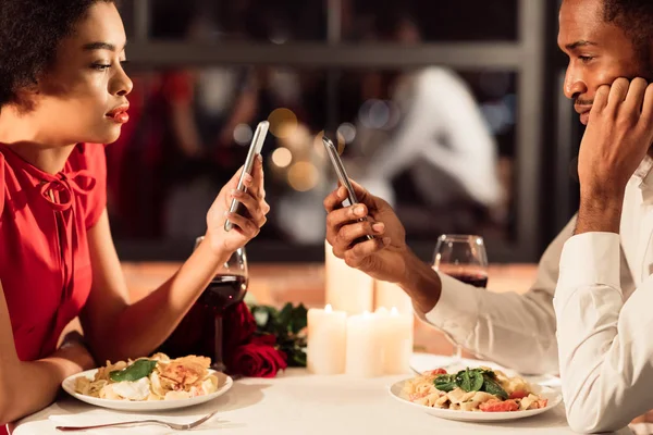 Uttråkad afroamerikanska par som använder telefoner under datum i restaurangen — Stockfoto