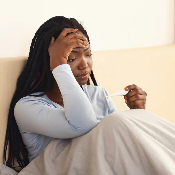Mujer negra sosteniendo termómetro teniendo fiebre y dolor de cabeza en el dormitorio —  Fotos de Stock