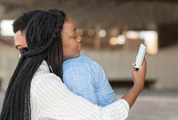 Черная женщина с помощью смартфона обнимается со своим парнем на улице — стоковое фото