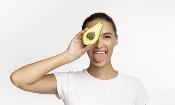 Woman Holding Avocado Half Covering Eye, Showing Tongue, White Background — Stockfoto