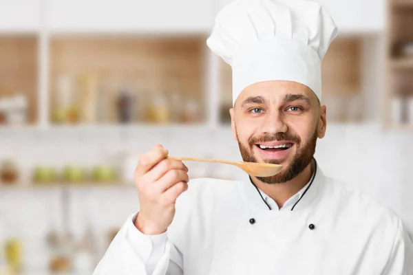 Веселый шеф-повар держит ложку дегустации еды на кухне в помещении — стоковое фото