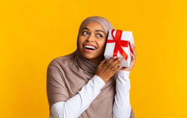 Joyful afro muslim woman holding present, shaking box, guessing whats inside