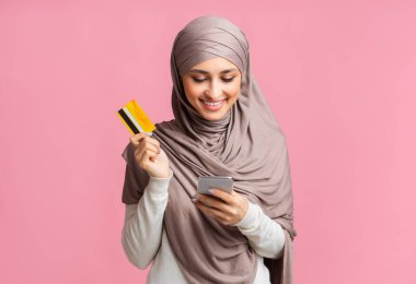 Happy muslim woman in headscarf using smartphone and holding credit card