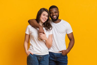 Happy multiracial couple hugging and posing over yellow background