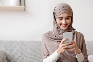Portrait of millennial muslim girl in hijab using smartphone at home