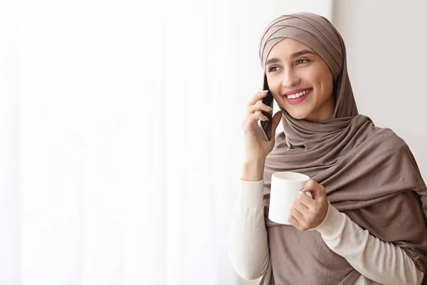 Boldog muszlim lány beszél mobilon és kávézik az ablak mellett — Stock Fotó