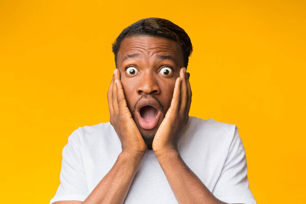 Удивлённый чёрный мужчина трогает лицо, позируя над оранжевым фоном
