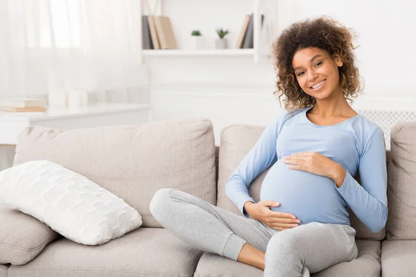 Feliz mujer embarazada sentada en el sofá y acariciando su vientre — Foto de Stock