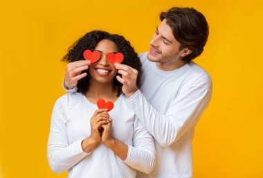 Sevgi dolu bir adam, kız arkadaşının gözlerini kırmızı kağıt kalplerle kapatıyor.