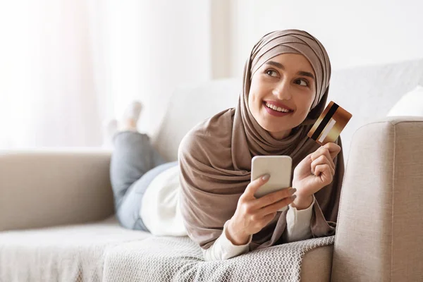 Pensive girl in hijab with smartphone and credit card lying on couch — Stock Photo, Image