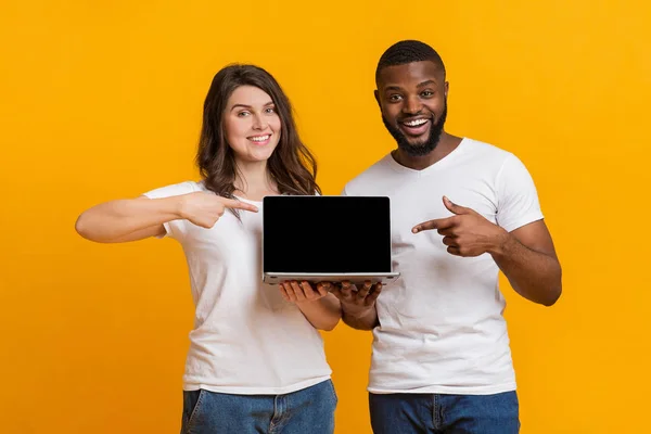 Alegre pareja multirracial sosteniendo y apuntando a la computadora portátil con pantalla negra — Foto de Stock