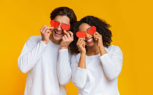 Romántica pareja interracial sosteniendo corazones rojos sobre los ojos y sonriendo — Foto de Stock