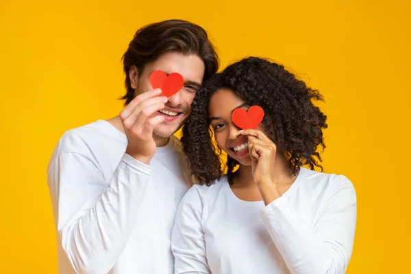 Romántica pareja interracial cubriendo los ojos con corazones de papel rojo en las manos — Foto de Stock