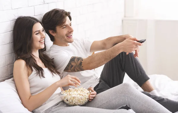 Millennial spouses watching tv with popcorn, sitting in bed — 스톡 사진