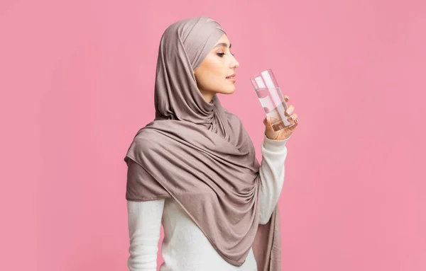 Wanita Muslim di kerudung minum air mineral bersih di atas latar belakang merah muda — Stok Foto