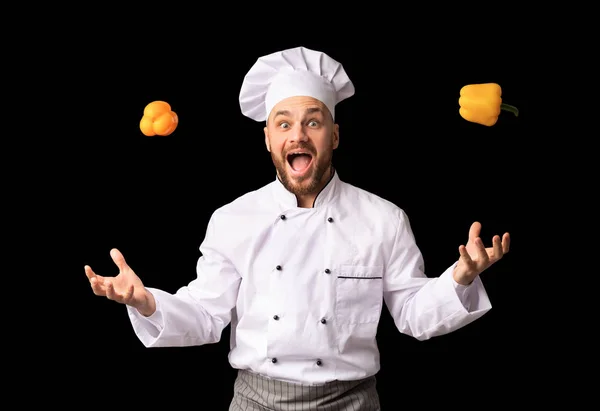 Engraçado Chef Man jogando pimentas amarelas em pé sobre fundo preto — Fotografia de Stock
