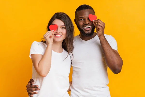 Smiling interracial couple covering eyes with red paper hearts — 스톡 사진