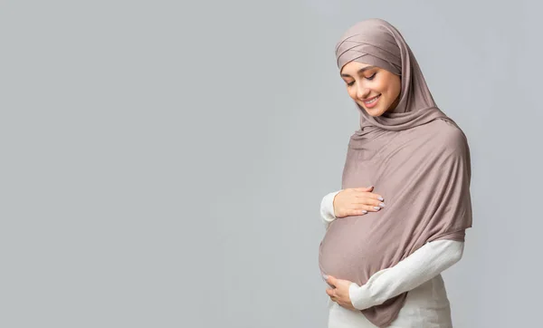 Wanita muslim hamil merangkul perutnya, berpose di atas latar belakang abu-abu — Stok Foto