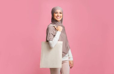 Başörtüsü takmış neşeli İslamcı kadın yanında alışveriş çantasıyla poz veriyor.