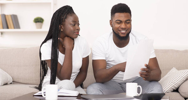 Улыбающаяся чёрная пара читает дома страховые документы, проверяет детали договора
