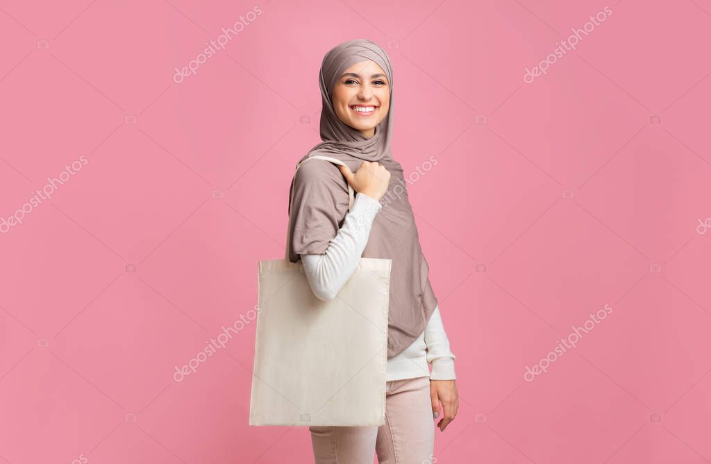 Eco shopping. Cheerful arabic girl in hijab posing with tote canvas shopper bag with place for mockup, pink background