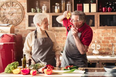 Neşeli kıdemli adam karısını eğlendiriyor ve mutfakta öğle yemeği hazırlıyor.